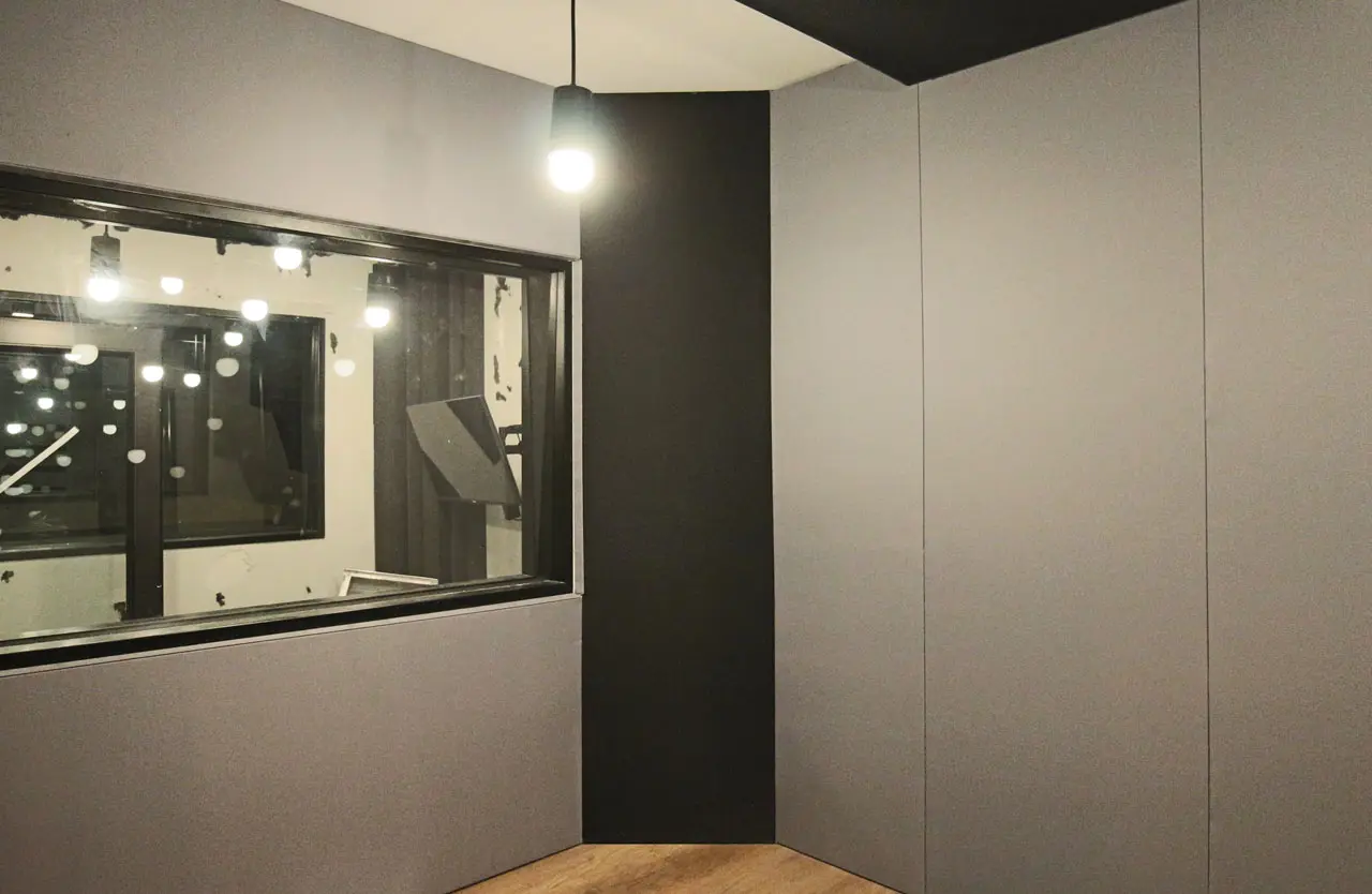 A modern, soundproof recording studio with gray walls, a large interior window looking into another room, and exposed light bulbs hanging from the ceiling. The floor is wood.