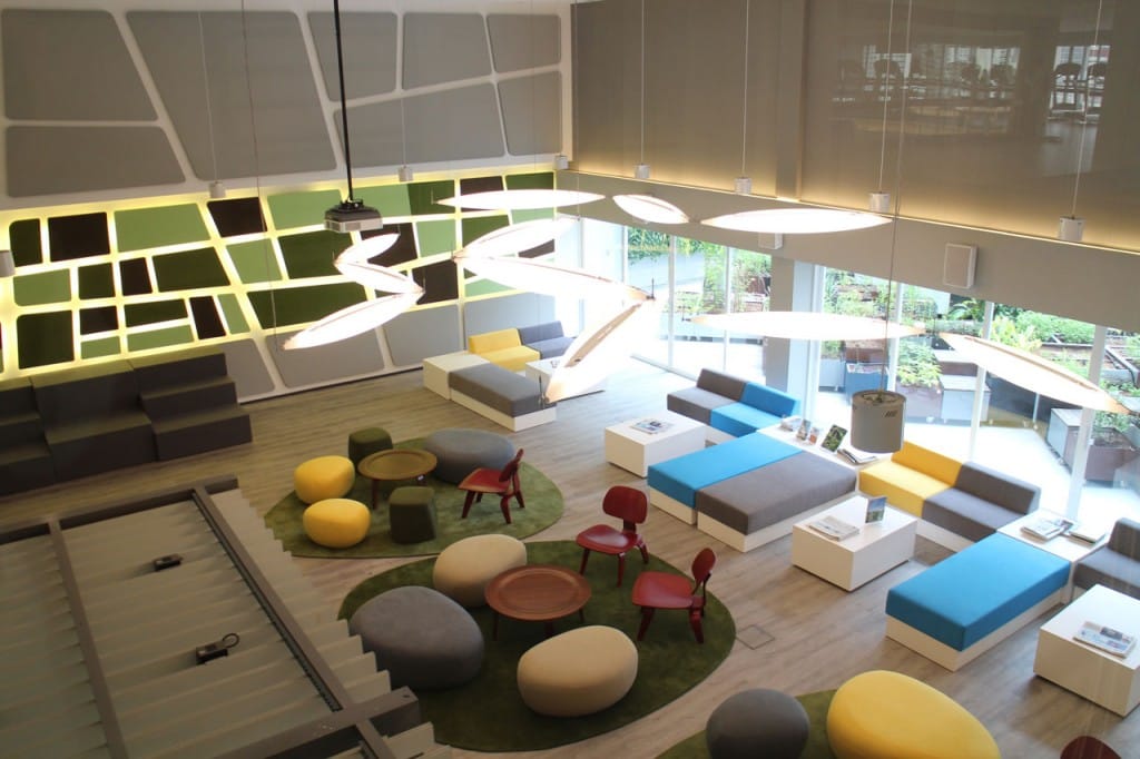 A modern, brightly lit lounge with geometric wall patterns, modular gray and blue seating, round ottomans, red chairs, green rugs, and unique angular light fixtures, with large windows overlooking greenery.