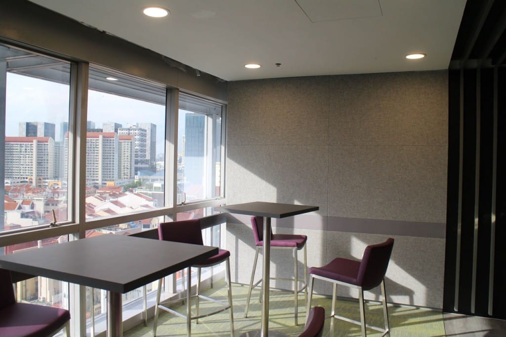A modern indoor seating area with tall tables and purple chairs by large windows, overlooking a cityscape of high-rise buildings under daylight. The walls are gray and the space is well-lit.