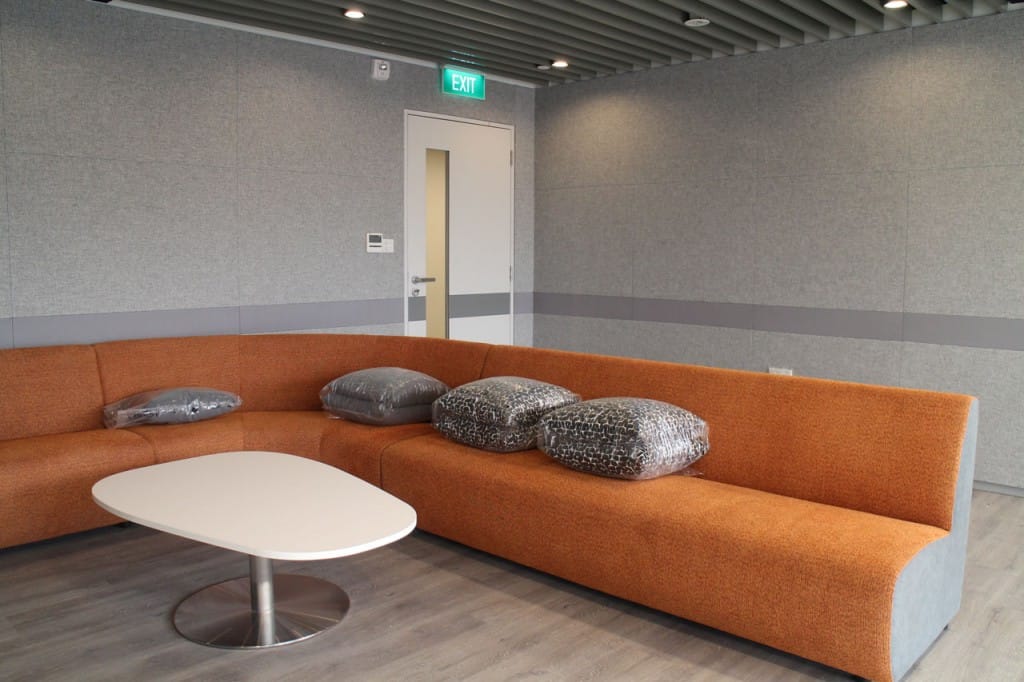 A modern lounge with an orange sectional sofa, several plastic-wrapped pillows, a white oval coffee table, and light gray walls and flooring. An emergency exit door is seen in the background.