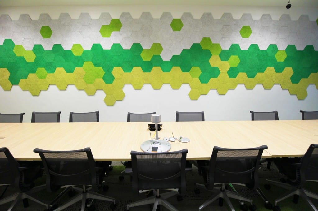 A modern conference room with a large wooden table, black mesh chairs, and a decorative wall featuring green, yellow, and gray hexagonal panels arranged in a honeycomb pattern.
