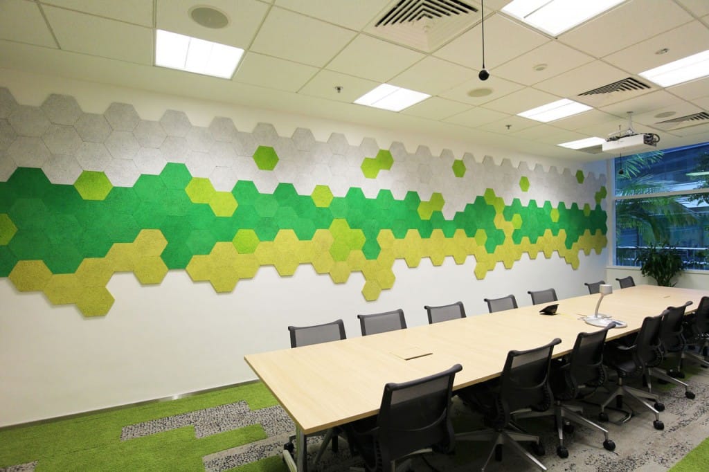 A modern conference room with a long wooden table, black office chairs, and a wall decorated with hexagonal panels in shades of green, yellow, and gray. Ceiling lights and a window with plants are visible.
