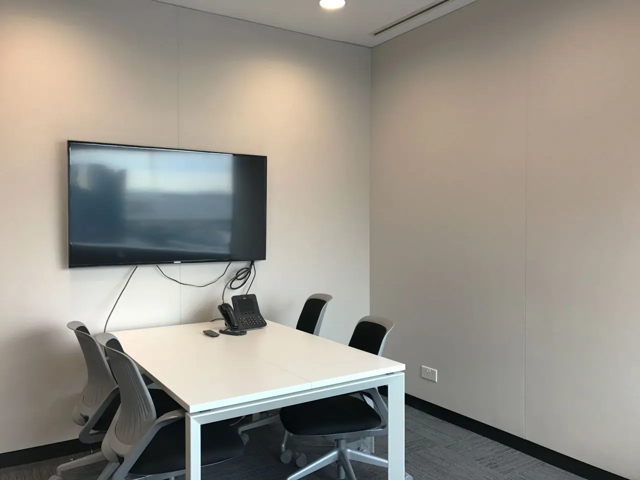 A small conference room with a white table, four office chairs, a wall-mounted flat-screen TV, and a telephone on the table. The room has light-colored walls and gray carpeted flooring.