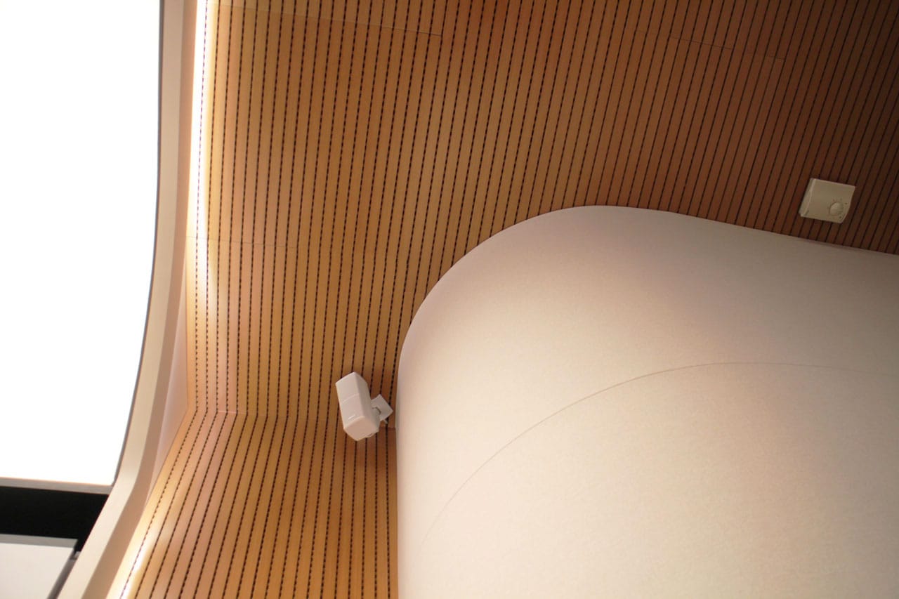 Curved wooden slat ceiling with a smooth, rounded white structure and a white security camera attached to the ceiling. Warm lighting accentuates the modern architectural design.