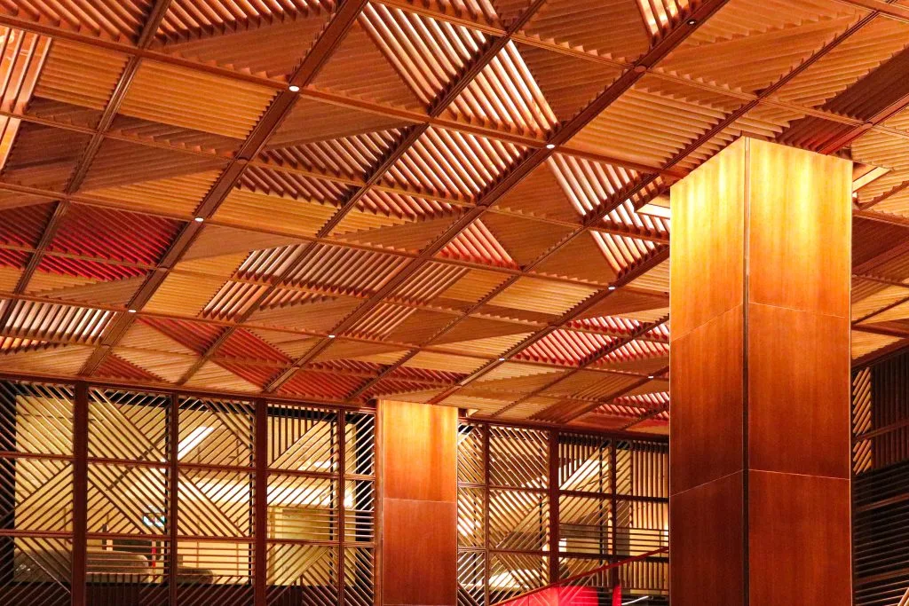 Interior view of a room with a ceiling and walls featuring geometric, diamond-shaped wood panels in warm tones. Two large wooden pillars extend from the floor to the ceiling, enhancing the modern, textured design.