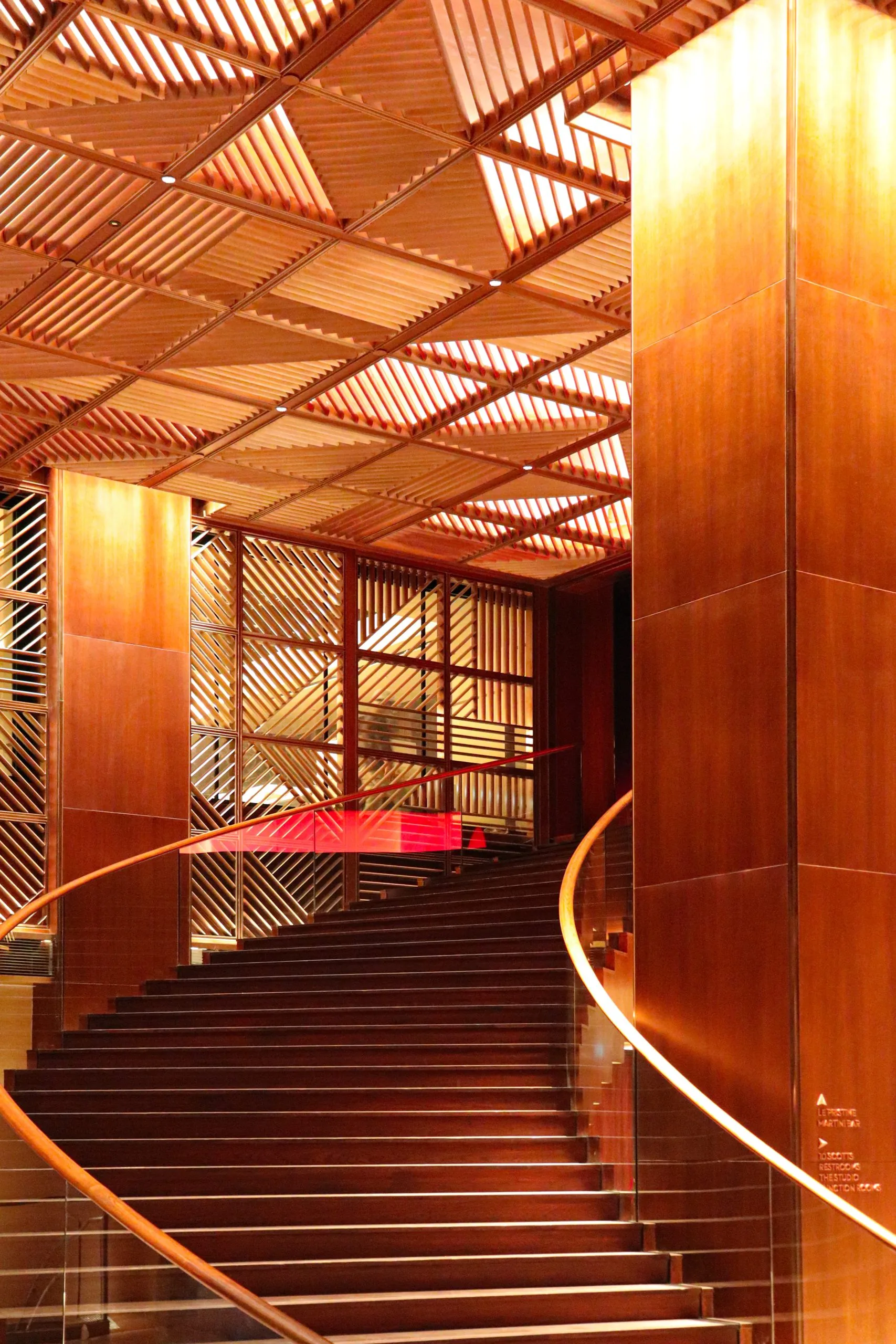 A grand curved staircase with wooden steps and railings, surrounded by tall wooden panels and a geometric wooden ceiling, illuminated by warm lighting.
