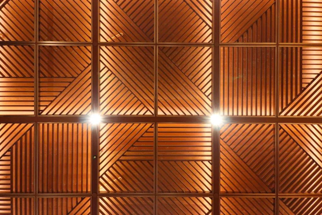 Geometric wooden ceiling with square panels featuring diagonal lines and two bright ceiling lights shining from the lower third of the pattern.