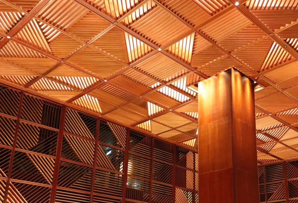 A modern interior with a geometric wooden ceiling and wall panels, featuring a central column. Warm lighting highlights the patterns and creates an inviting atmosphere.