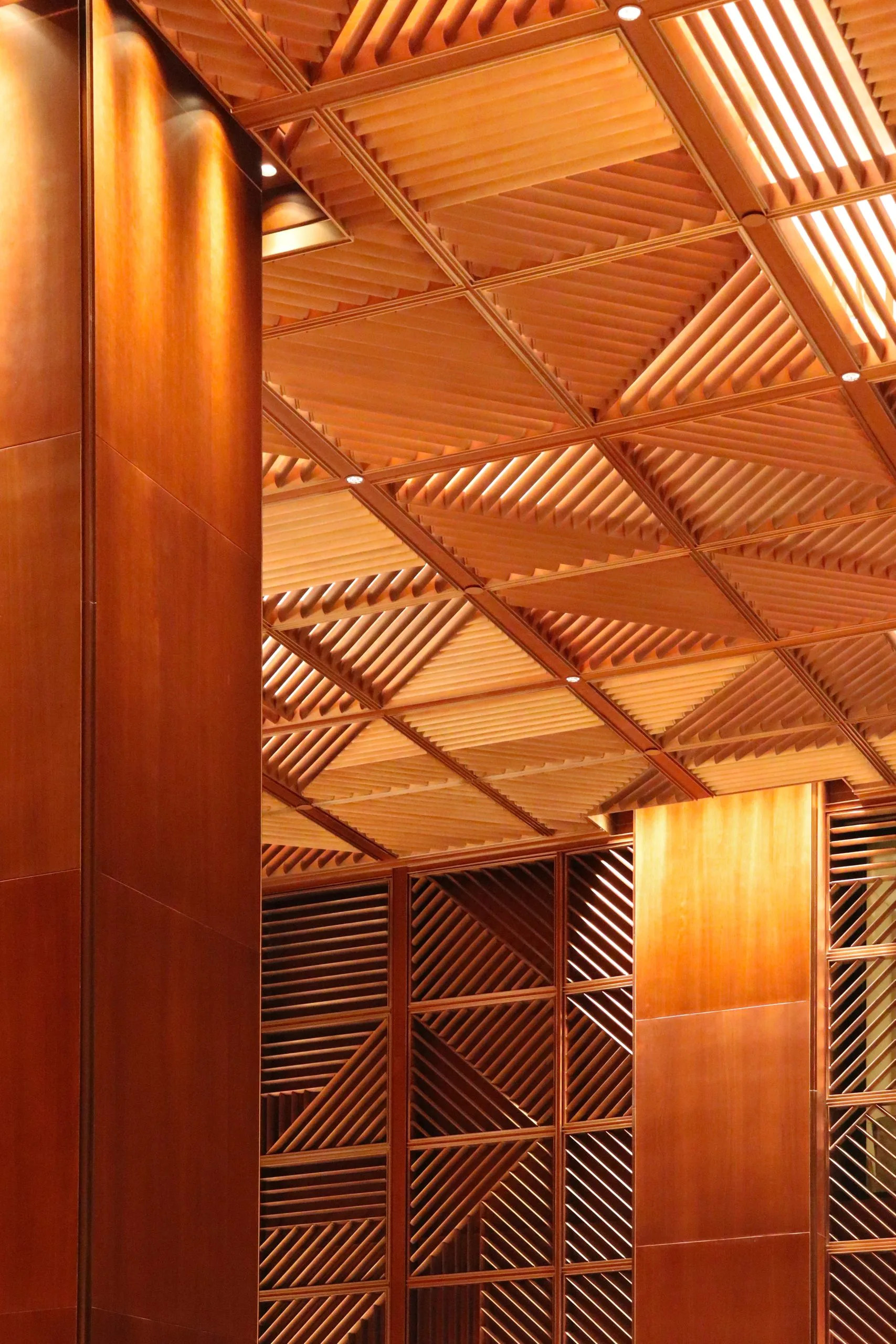 A close-up view of an interior with warm wooden panels and geometric patterns on the walls and ceiling, illuminated by soft, recessed lighting. The intricate design creates a modern, elegant atmosphere.