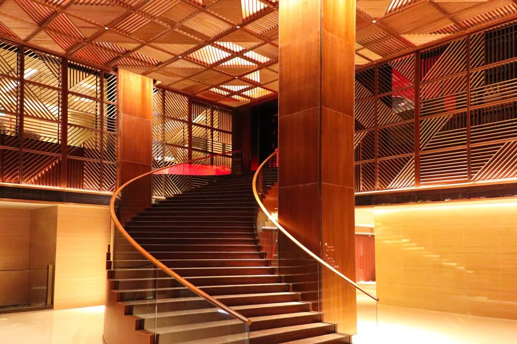 A grand, curved staircase with glass railings and wood accents leads up to a mezzanine in a modern interior, featuring warm lighting and decorative wooden paneling on the walls and ceiling.