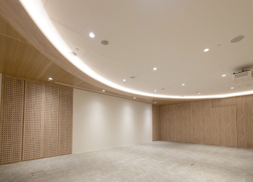 A spacious, empty room with light wood paneling on the walls, a curved white ceiling with recessed lighting, and a blank white wall; the floor appears to be bare concrete.