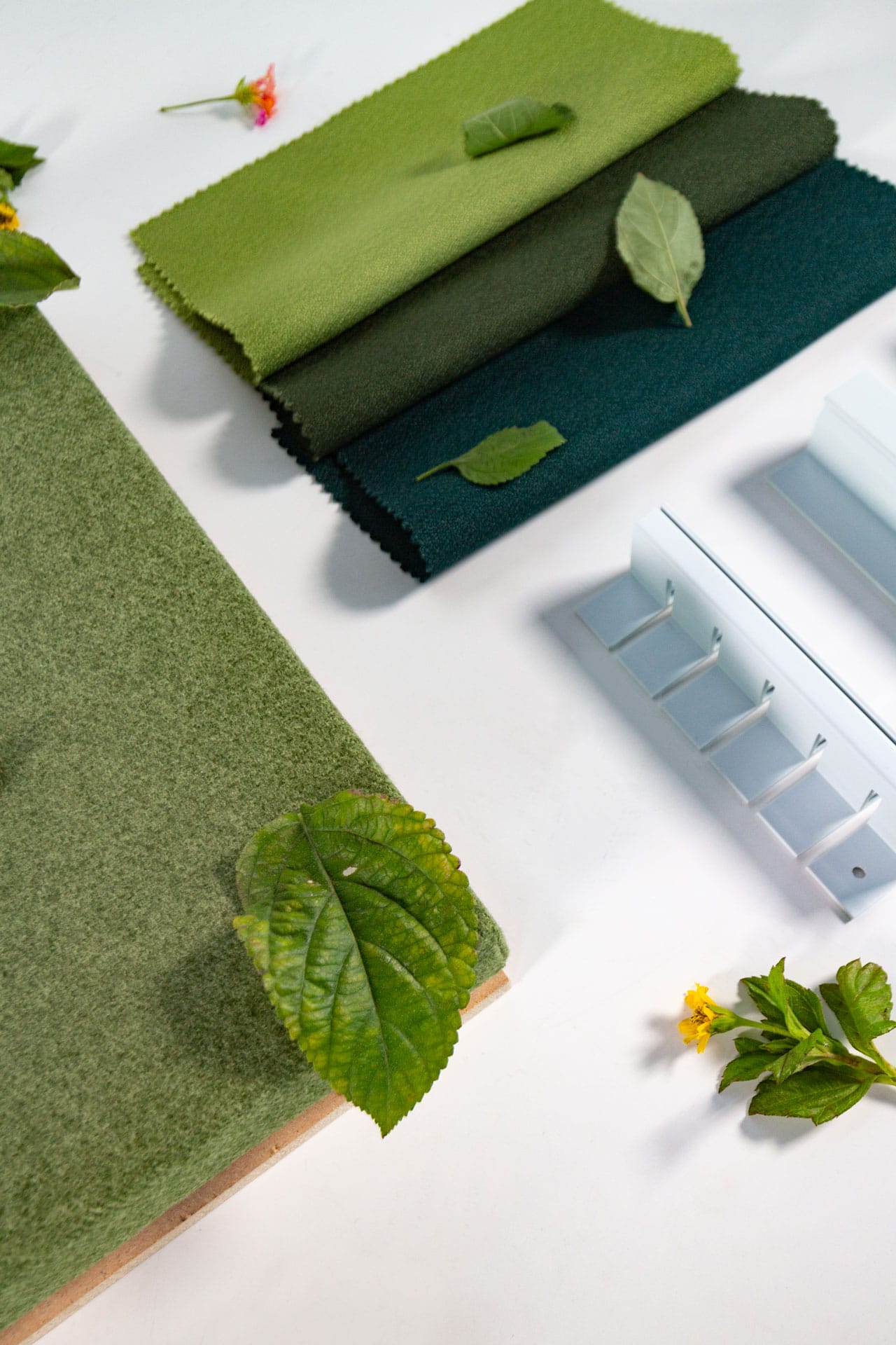Green and teal fabric swatches, a green textured board, some leaves, and small yellow flowers are arranged on a white surface next to a white wall hook rail.