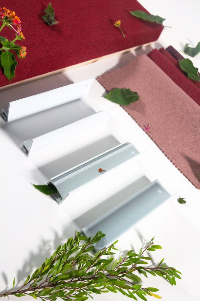 Metal brackets arranged on a white surface with fabric swatches, green leaves, small flowers, and plant stems scattered around them. The scene has a mix of industrial and natural elements.