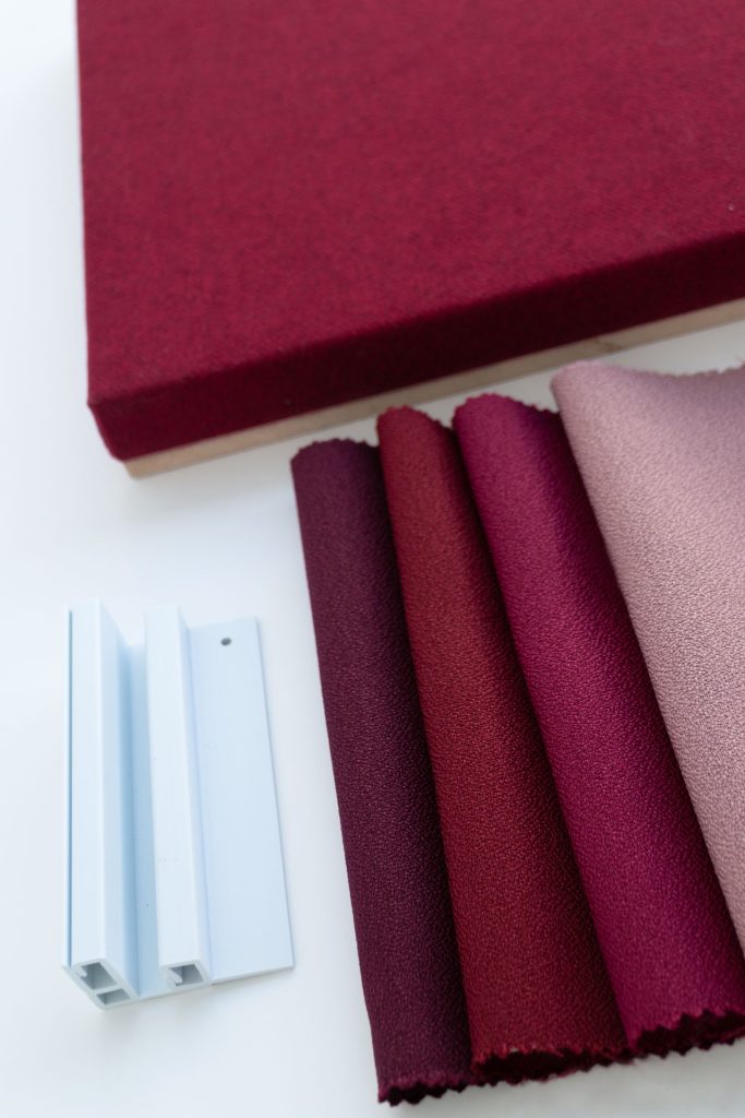 A selection of red, burgundy, and pink fabric swatches is arranged next to a matching fabric-covered board and two white plastic frame samples on a white surface.