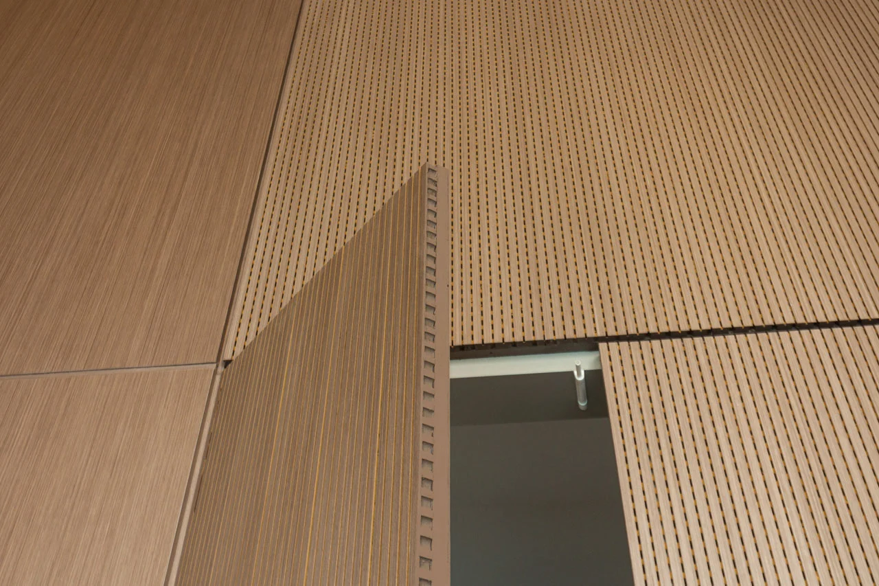 A close-up view of a modern wooden door, partially open, set within a wall of matching vertical wooden panels with small perforations. The door aligns seamlessly with the wall when closed.