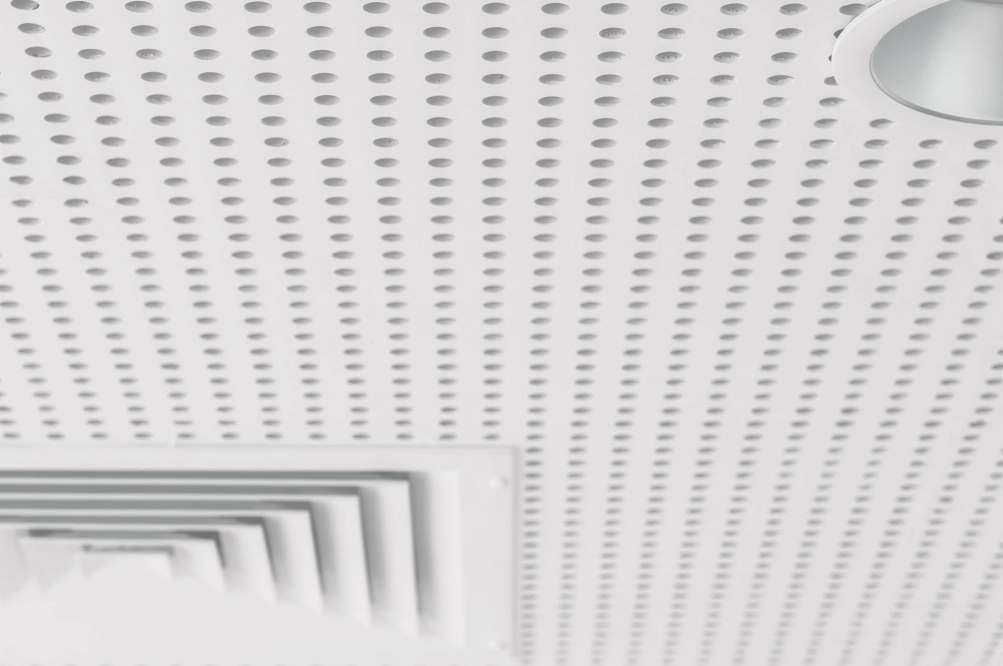 White perforated ceiling with circular holes, a recessed light fixture in the upper right corner, and a square air vent with layered slats in the lower left corner. The overall appearance is clean and minimalistic.
