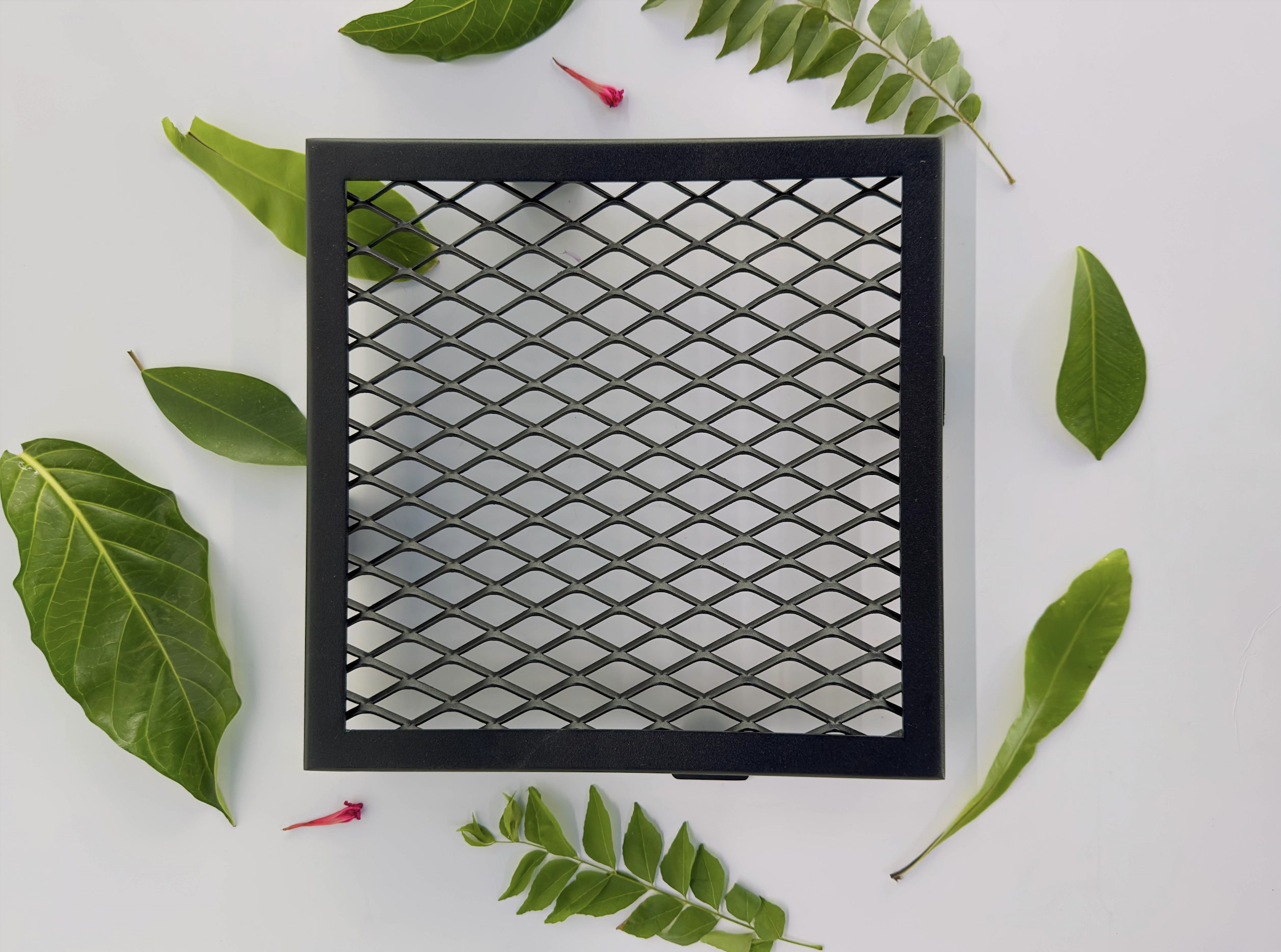 A black, square metal mesh panel is placed on a white surface, surrounded by various green leaves and a small red flower. The mesh has a diamond-shaped grid pattern.