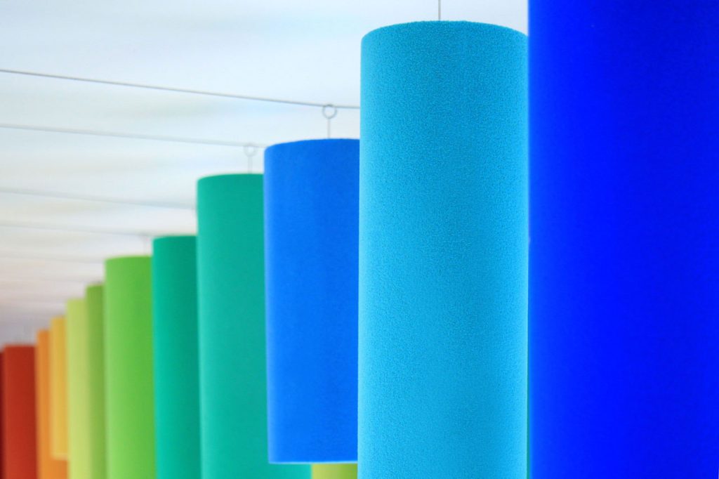 Colorful cylindrical columns hang from the ceiling in a row, arranged in a gradient from red, orange, yellow, and green to blue, creating a vibrant and abstract geometric pattern.