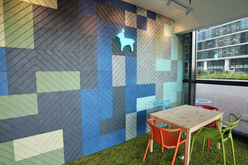 A small table with four colorful chairs sits on green carpet next to a window. The wall is covered in geometric blue and green panels with a glowing animal silhouette decoration.