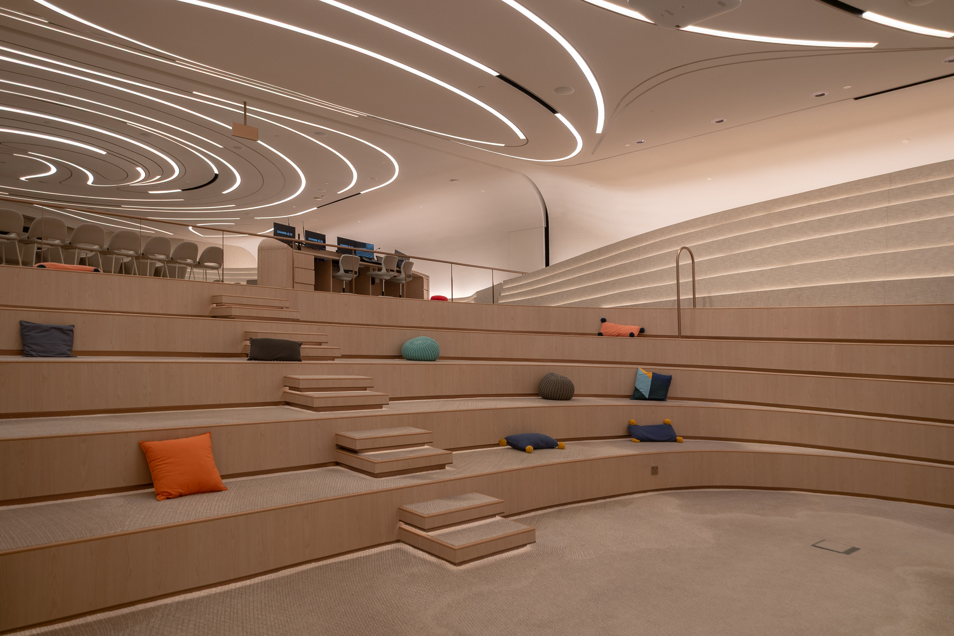Modern, curved auditorium with wooden tiered seating, scattered colorful pillows, steps leading up, and a wavy, illuminated ceiling design. A control desk with monitors sits at the back near empty chairs.