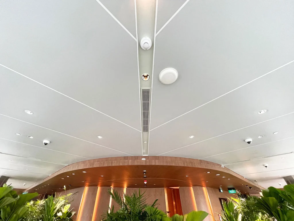 Ceiling with modern lighting, security cameras, and air vents in a contemporary indoor space. Below, green leafy plants and curved wooden architectural elements are visible.