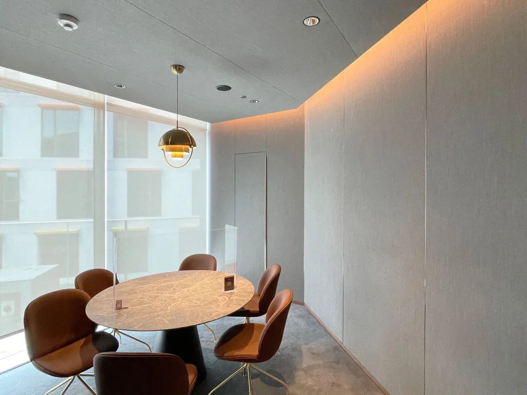 A modern meeting room with a round marble table, five brown chairs, a gold pendant light, large floor-to-ceiling windows, and minimalistic gray walls with subtle warm lighting.