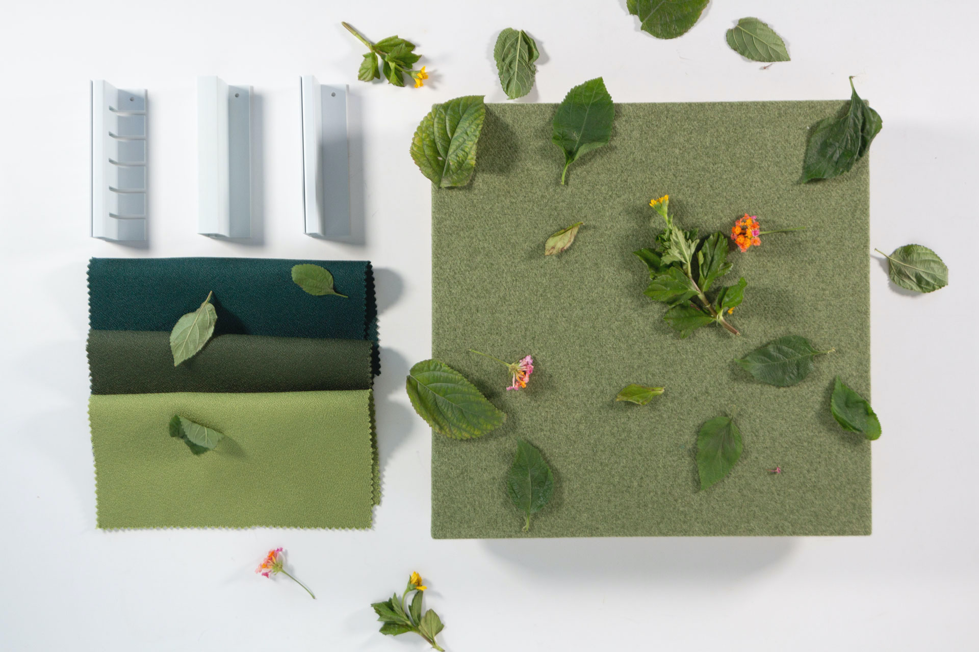 A green fabric square, scattered with leaves and small flowers, sits beside three rectangular gray samples and two fabric swatches—one dark green, one light green—on a white background.