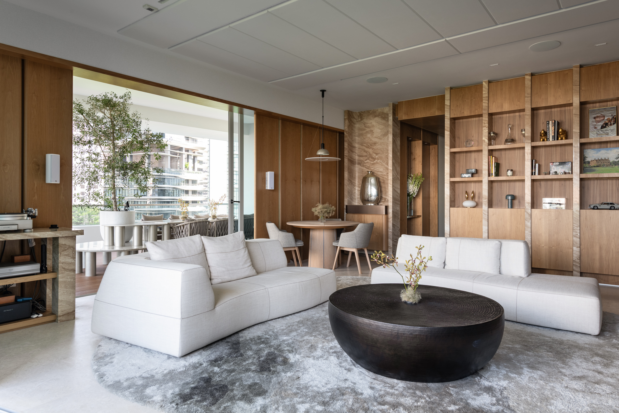 Modern living room with light wood paneling, white sectional sofas, a round dark coffee table, and shelves with decor. Large windows and an open dining area create a bright, airy atmosphere.