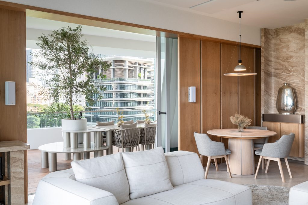 Modern living and dining area with neutral tones, light wood accents, a round dining table with four chairs, a white sofa, sliding glass doors, potted plant, and a balcony overlooking contemporary buildings.