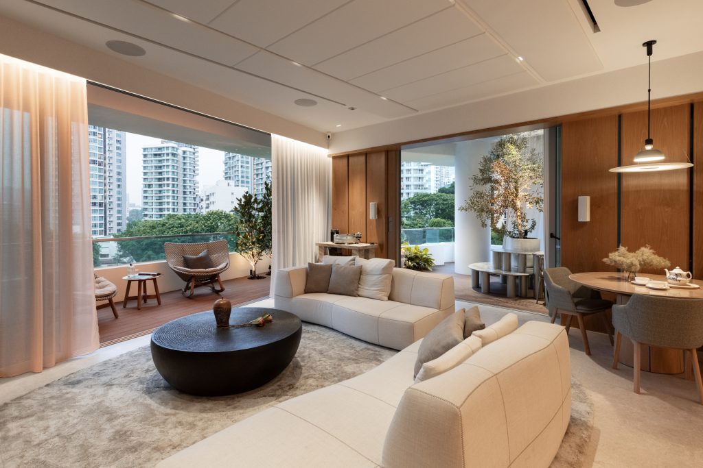 A modern living and dining area with light neutral furnishings, a large curved sofa, round black coffee table, sliding doors opening to a balcony, and city buildings visible outside. Warm lighting creates a cozy atmosphere.