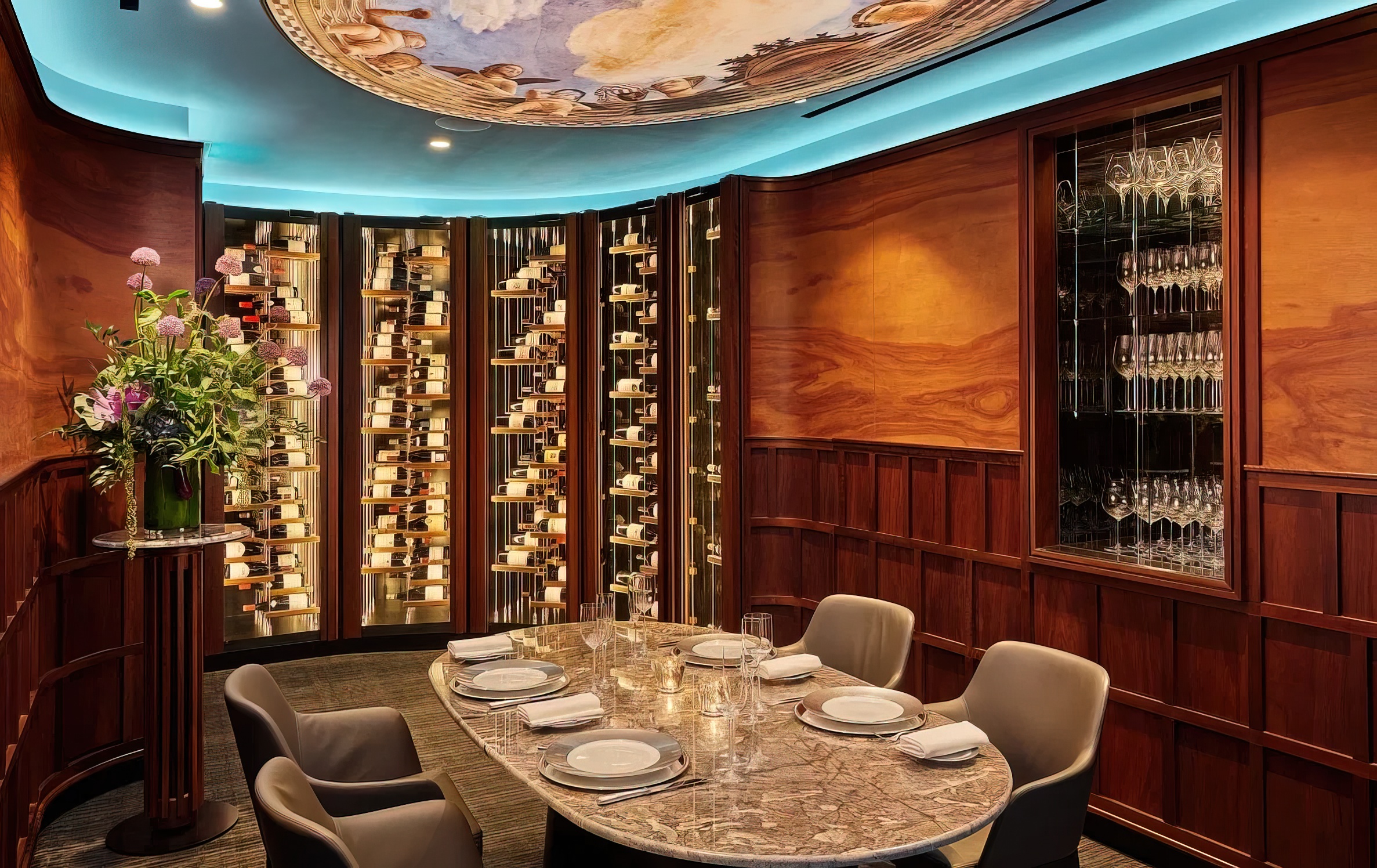 A luxurious private dining room with a marble table set for six, plush chairs, a wall of wine bottles, wood-paneled walls, a glass cabinet of stemware, and a ceiling mural under blue accent lighting.