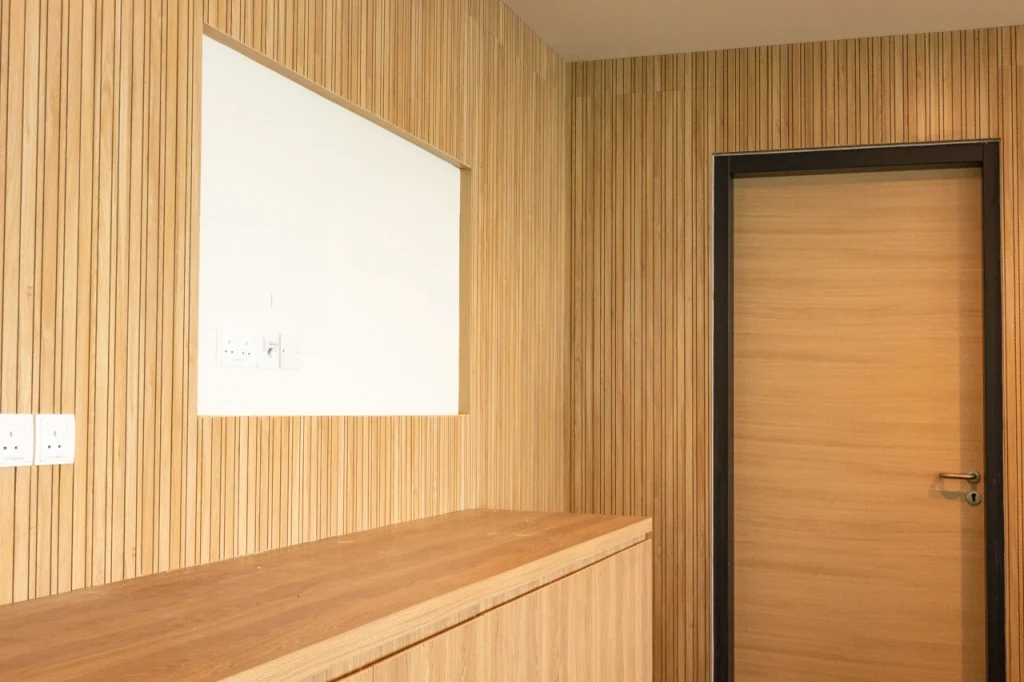 A modern interior with light wood paneling on the walls, a wooden cabinet, a door with a dark frame, several electrical outlets, and a large rectangular cutout above the cabinet.