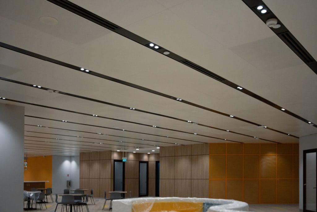 A modern, spacious room with a white ceiling featuring recessed lighting, light wood-paneled and yellow accent walls, gray chairs, and tables. Some furniture is wrapped in plastic, suggesting recent renovations.