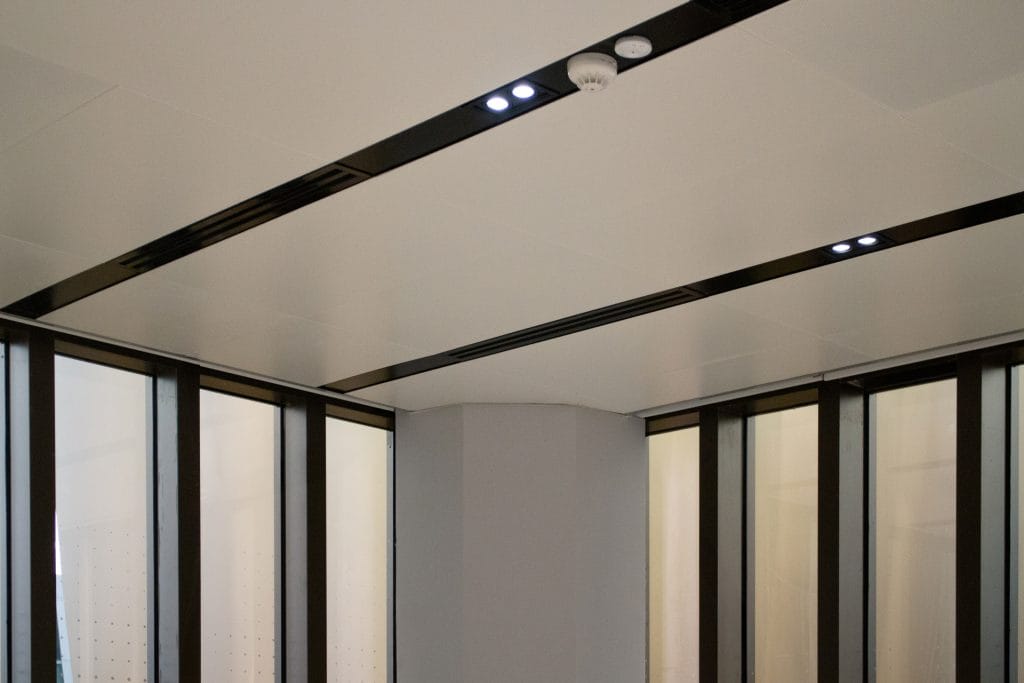 A modern indoor ceiling with recessed lighting, black trim, and a smoke detector. Large glass windows with dark frames line the walls, letting in natural light. The overall design is minimalistic and clean.