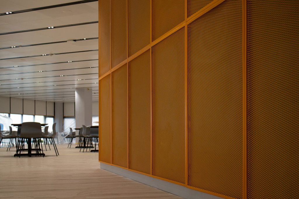 A modern interior features a large orange perforated wall divider on the right and a spacious seating area with gray chairs and tables on a light wooden floor in the background.