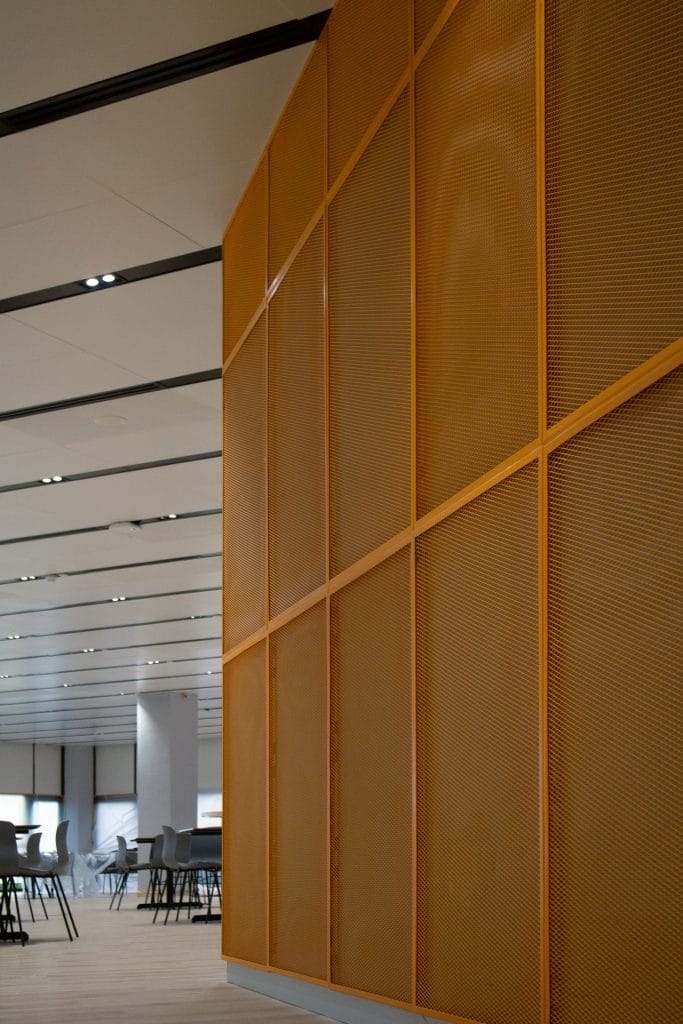 A modern interior space featuring a large orange perforated metal wall on the right; in the background, minimalist tables and chairs are arranged in a well-lit open area with ceiling lights.