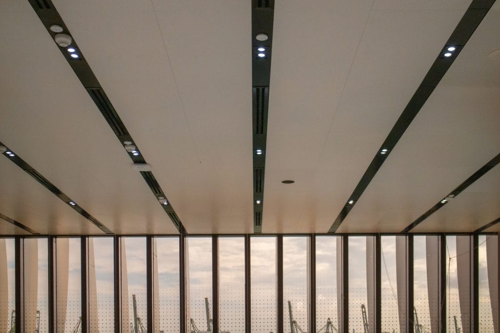 A modern ceiling with linear recessed lighting and panels, above large floor-to-ceiling windows that reveal a hazy sky and faint outlines of structures outside.