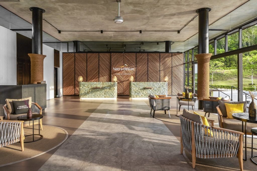 Spacious hotel lobby with modern decor, featuring a wooden reception desk, wicker chairs with yellow cushions, large windows letting in natural light, and greenery visible outside. The wall behind the desk displays Natra Bintan.