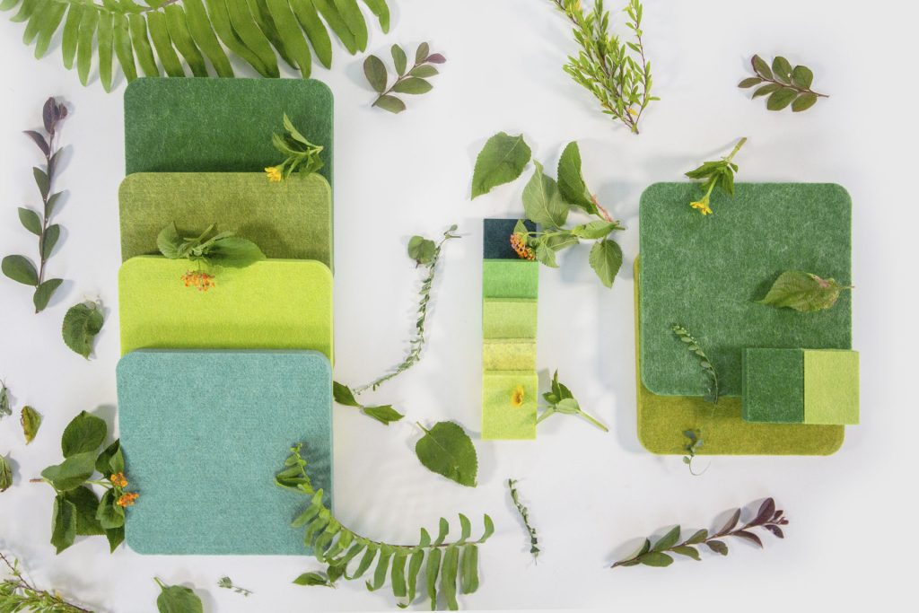 Assorted green fabric swatches and small color sample cards are arranged on a white surface, surrounded by various green leaves and small plant stems.