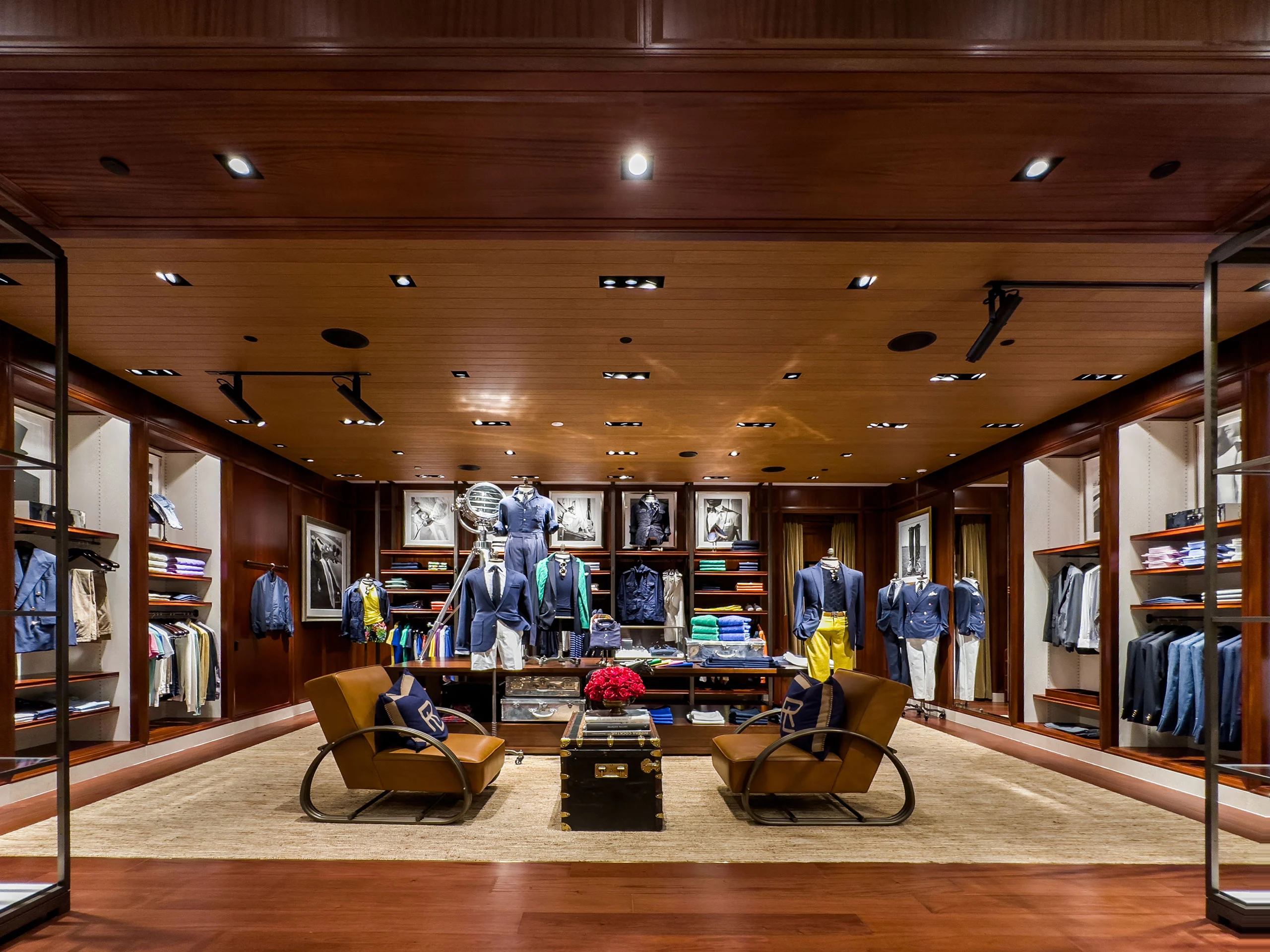 A stylish, upscale clothing store with wooden walls and ceiling, shelves of neatly folded clothes, mannequins dressed in casual wear, leather chairs, and a central display table with red flowers.