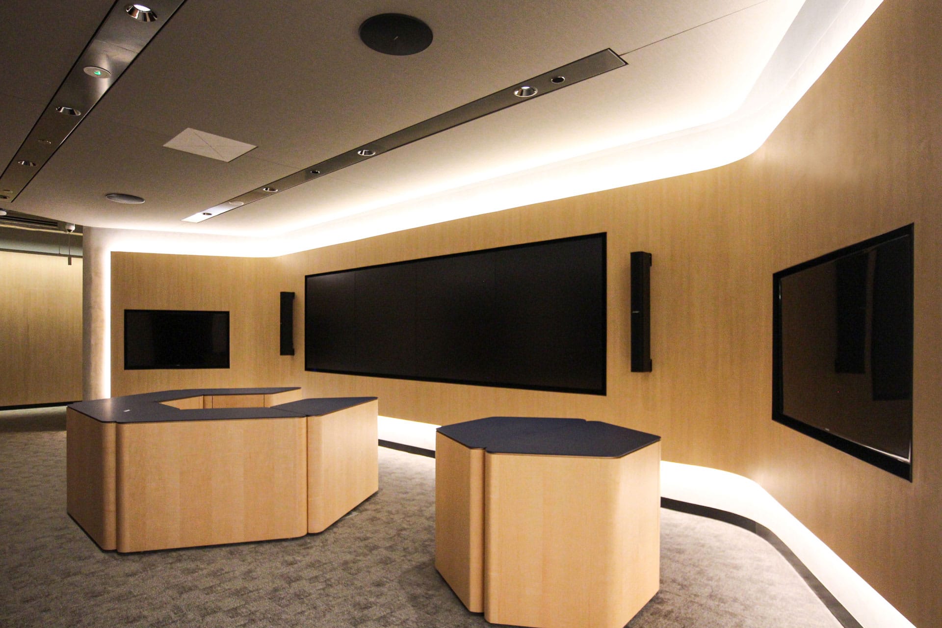 A modern conference room with light wood walls, large black screens on the walls, two polygonal tables in the center, and soft recessed lighting creating a sleek, minimalist atmosphere.