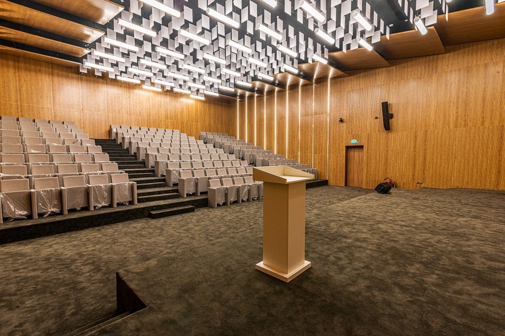 A modern, empty auditorium with tiered seating, a central podium on a carpeted stage, wooden walls, and a ceiling decorated with geometric light panels.