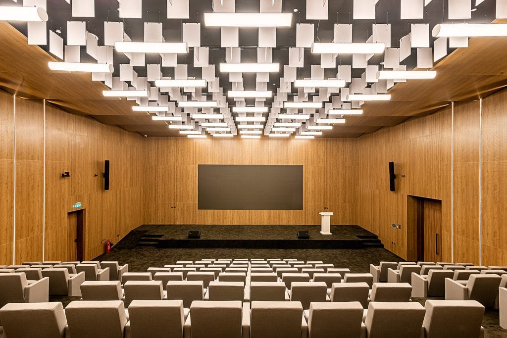 A modern auditorium with wooden walls, rows of gray seats facing a stage with a podium and large screen, and a ceiling featuring geometric light fixtures.
