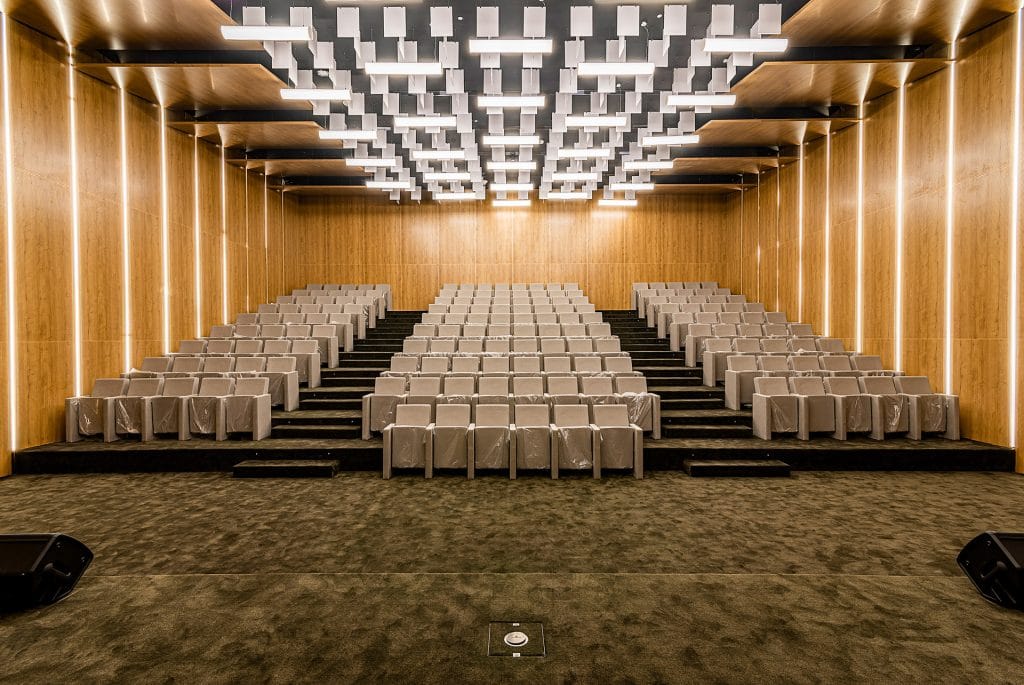 A modern lecture hall with tiered rows of grey seats, wooden paneled walls, and a unique geometric ceiling with bright lights. The seats are unoccupied and covered in plastic.