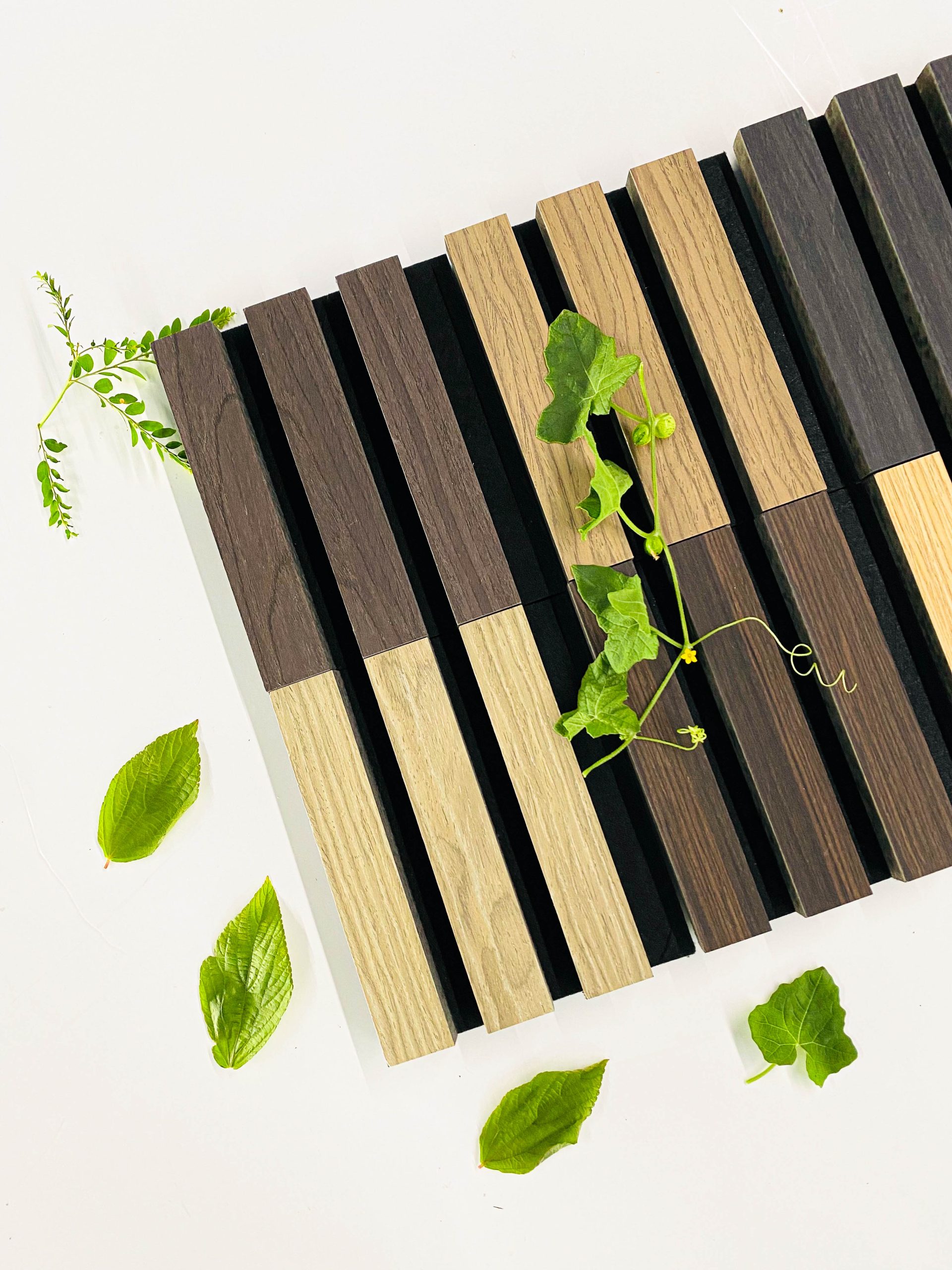 A pattern of alternating dark and light wooden slats arranged vertically, with green leaves and vines placed on and around the wood on a white surface.