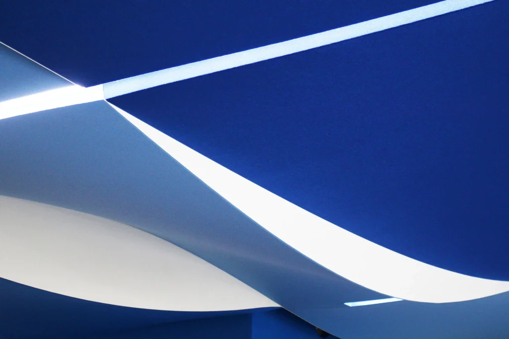 Abstract view of a ceiling with curved blue and white panels intersected by a thin, bright light, creating a modern and artistic design.