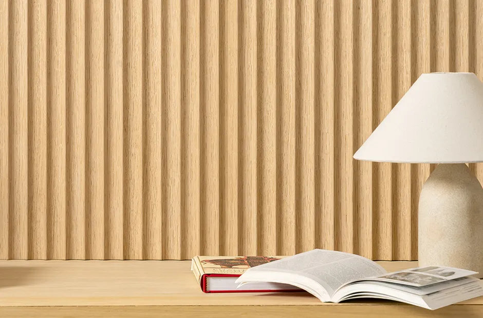 A minimalist desk with an open book, a closed book, and a beige table lamp sits in front of a textured wooden panel wall.