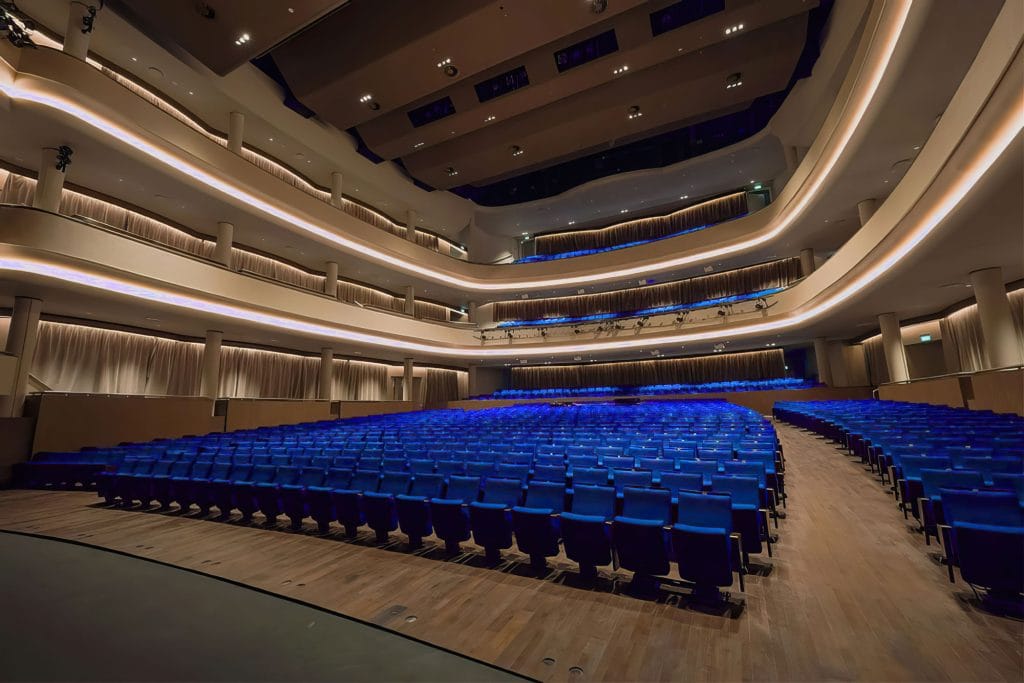 A modern, empty theater auditorium with rows of blue seats, wooden flooring, and three tiers of balconies, all illuminated by soft, yellow lighting.