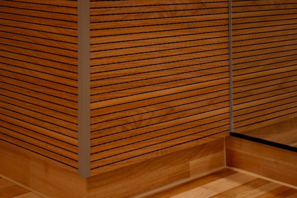 Close-up of a wooden wall with horizontal slats, showing detailed wood grain patterns. There is a metal strip and a glass panel at the corner, reflecting part of the wooden surface and floor.