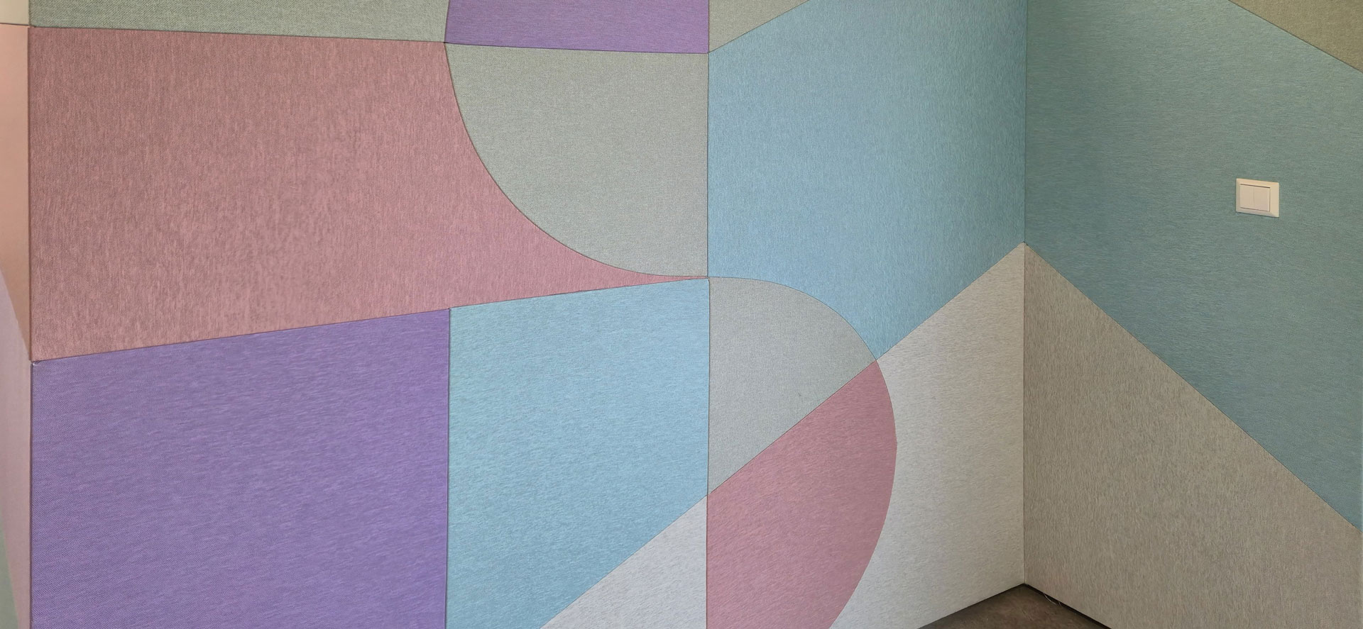 Geometric wall art with pastel-colored panels in shades of blue, purple, pink, and gray, forming intersecting shapes and lines. A white electrical switch is visible on the right side of the wall.